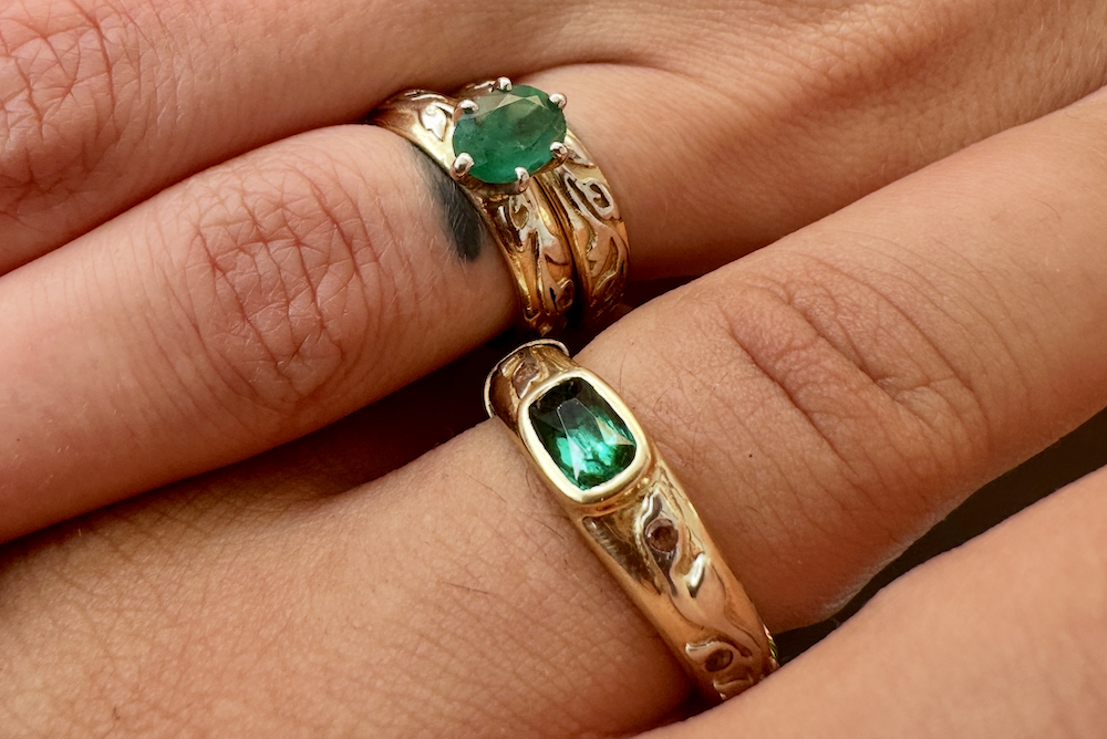 Three similar rings, each two-tone with an 18K yellow gold base and 14K white gold Flores motif by Reflective Jewelry. One ring is set with an oval emerald in a prong setting, another is set with an emerald-cut emerald in a bezel setting, the third does not contain a stone.